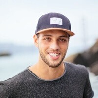 Man wearing a navy cap and dark T-shirt, smiling outdoors by the water in the background.