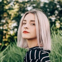 Portrait of a young woman with light lavender hair looking over her shoulder, outdoors among greenery and plants.